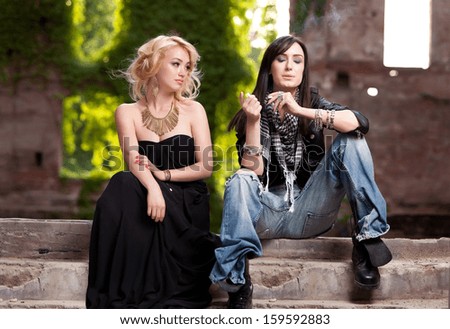 Strong contrast between very elegant and blonde woman sitting together with a daring brunette in torn jeans, lighting her cigarette.