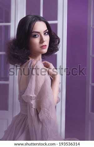 Young woman in a silk dress sitting in front of a large window doors.