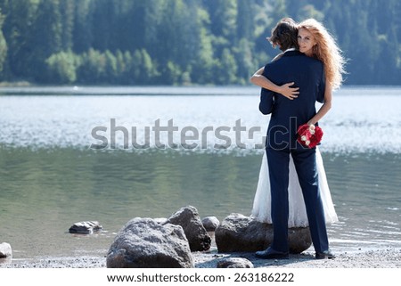 Beautiful married couple having a romantic moment near a lake. The bride stands on a big rock just to give him a lovely hug.