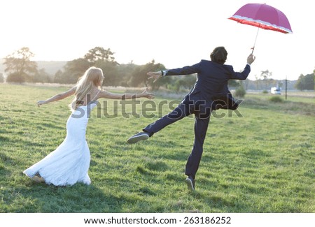 Married couple having fun on a field. The groom is trying to fly away using an umbrella while the bride is trying to catch him.