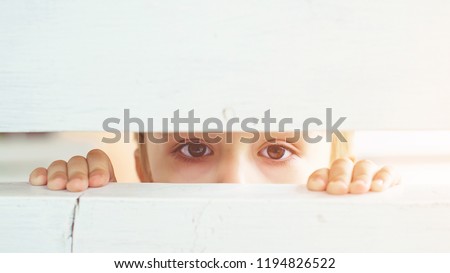 Frightened child is spying through a wooden fence. Scared child boy. Human emotion, facial expression. Sad kid outdoors. Bored little boy