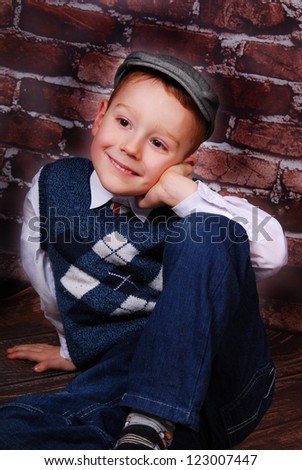 elegant little boy in golfer cap and wool vest sitting on a brick wall background