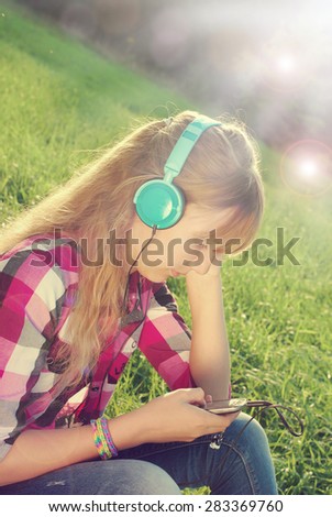 young beautiful girl with headphones on ears listening music in smartphone on the meadow in vintage style