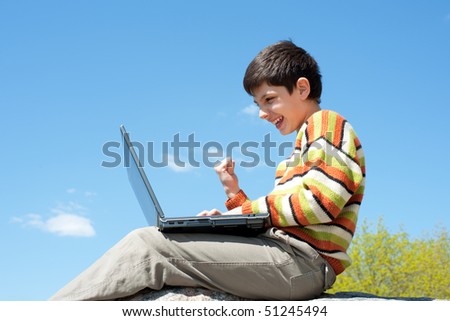 A shouting boy is sitting on the rock outside and playing a computer game