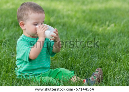 A little toddler in green is drinking milk from a glass sitting on the green grass in the park