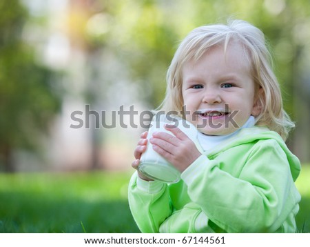 A laughing little girl in green is drinking milk from a glass in the park