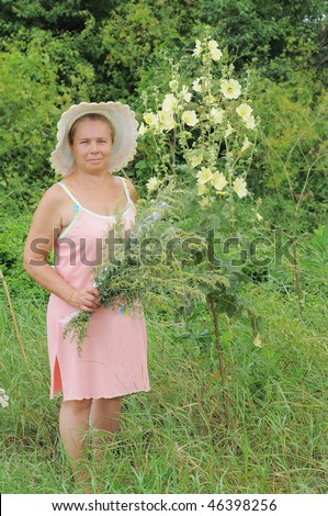 The elderly woman in a summer hat on the fringe of the forest and wildgrowing flowers