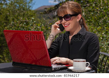 Business woman with sunglasses on sitting at an outdoor table with her laptop and cell phone smiling.