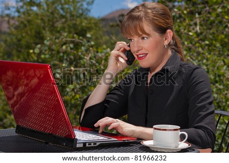 Business woman sitting at an outdoor table talking on her cell phone and typing on her laptop.