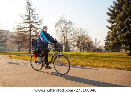 elderly man wearing glasses, a blue jacket, black trousers and a gray hat, riding a bike on the sidewalk