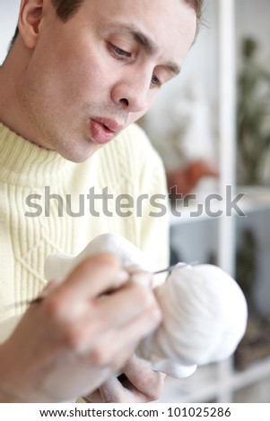 Sculptor carefully works on plaster statuette with sculptor tools and blows off plaster crumbs.