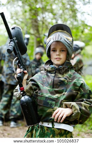 Boy in the camouflage suit holds a paintball gun barrel up , standing on the paintball ground with group of players on the background.