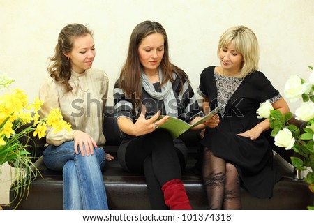 three young women sit on black leather couch and browse journal