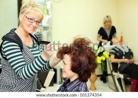 Smiling hairdresser makes hair styling for woman by rake-comb and looks at camera in beauty salon; focus on barber; other barber washes second client head