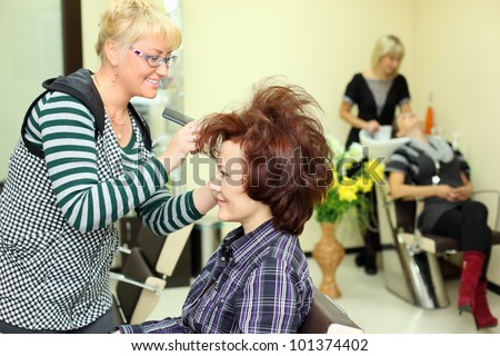 Smiling hairdresser makes hair styling for woman by rake-comb in beauty salon; focus on client; other barber washes second client head