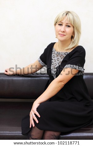 blonde woman dressed in black dress sits on black leather couch