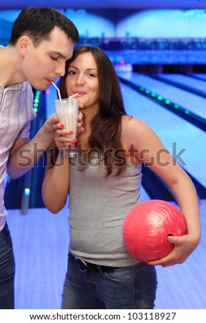 Woman and man together drink milk cocktail from one glass in bowling; woman holds red ball; focus on woman