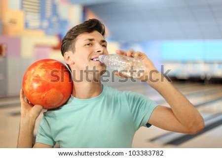 Happy man holds orange ball and drinks water from bottle in bowling club; shallow depth of field