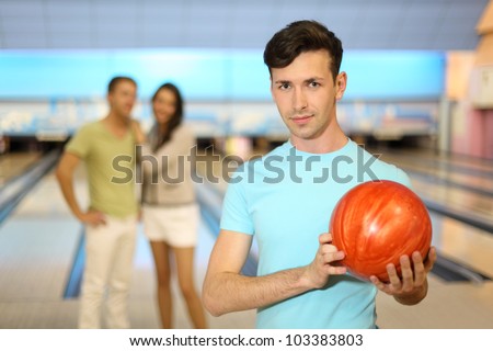 Young man with orange ball; couple stands behind him in bowling club; focus on right man; shallow depth of field