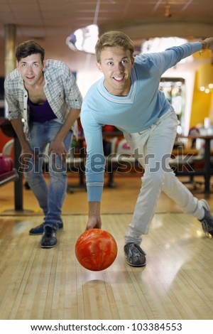 Young smiling man throws ball in bowling; friend looks at aim; shallow depth of field