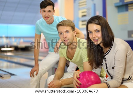 Two men and woman sit in bowling club; woman holds pink ball; focus on center man; shallow depth of field