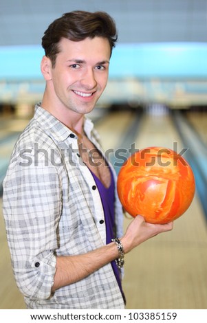 Young happy man wearing in white checkered shirt holds orange ball in bowling club; shallow depth of field