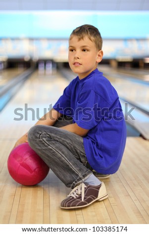 Boy dressed in blue T-shirt with pink ball sits on floor in bowling club; shallow depth of field; full body