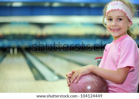Little girl dressed in pink T-shirt sits and holds ball in bowling club