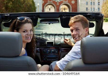 Young couple, looking back, sits in a cabriolet against building with two arches, girl is on a driver seat.