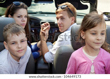 Young couple sits in cabriolet on front seats looking back, two children sit on  backseats, back view