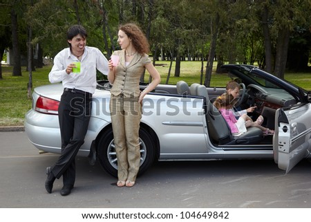 Parents stand near gig, talk and drink something from plastic cups, children play in the car