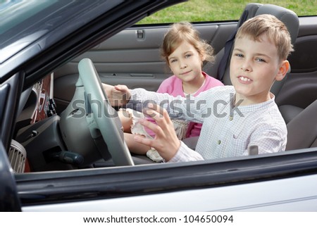 A boy sits on a driver seat in the open top car and his sister sits on a passenger seat, both look into a camera, close-up.