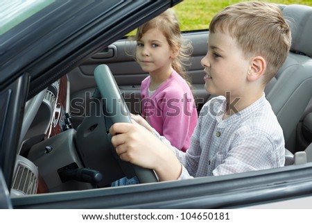 Boy sits on a driver seat in open top car and his sister sits on passenger seat, focus on boy, close-up.