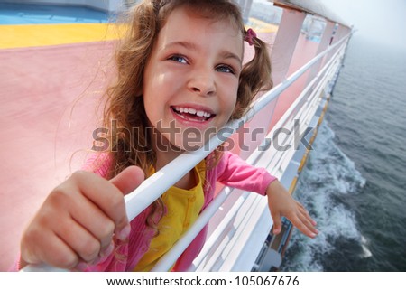 Little beautiful girl stands on board of large ship, clings to railing and laughs