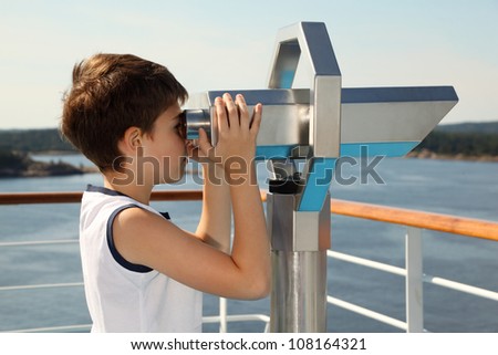 Boy stands on board of ship and looks through binoculars at beautiful landscape