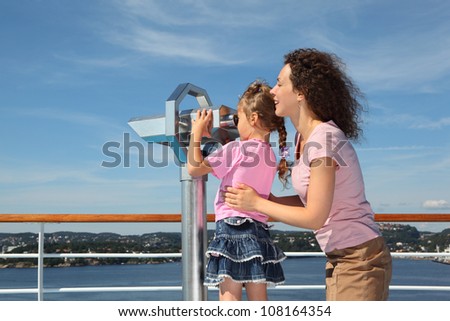 Mother and daughter stand on deck of ship; girl looks through binoculars at landscape