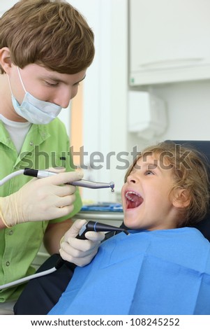 Young dentist holds saliva suction and grinding drill, happy girl opens her mouth in dental clinic.