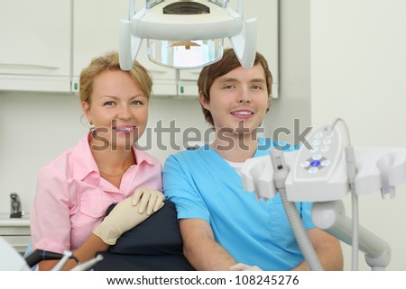 Two smiling dentists sit in cabinet of dental clinic near chair for patients.