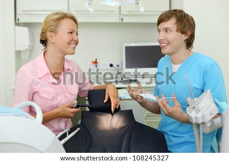 Two smiling dentists sits in cabinet of dental clinic and look at other before work.