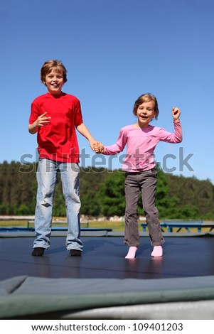 Smiling brother and sister jump on trampoline on sunny summer day