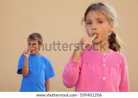 Girl and boy suck multicolored candies. Sister looks at camera, brother stands behind her. Focus on girl.
