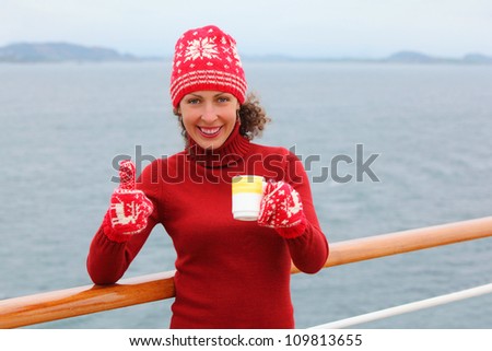 Woman wearing in red hat and mittens holds cup of hot tea and thumbs up at ship deck
