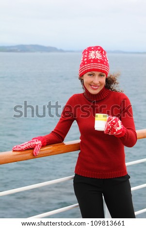 Smiling woman wearing in red hat and mittens holds cup of hot tea at ship deck