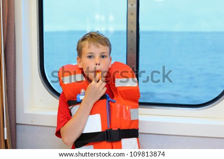 Boy dressed in life jacket blows whistle and stands near window in cabin of ship