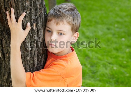 Portrait of boy in orange t-shirt, that stands, embracing tree in park