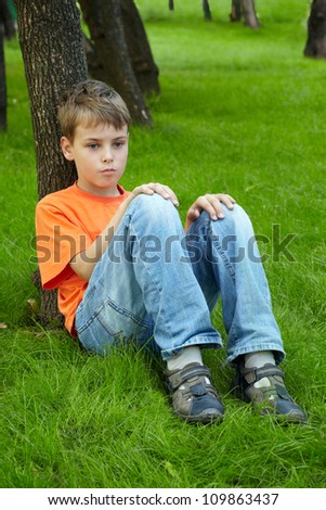 Boy in orange t-shirt sits with thoughtful face on grass, looking in front of himself and leaning his back on trunk of tree