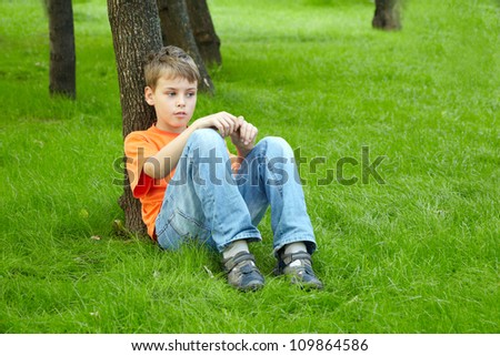 Boy in orange t-shirt sits with thoughtful face on grass, leaning his back on trunk of tree