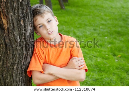 Portrait of boy in orange t-shirt with folded arms, standing leaned to tree  in park