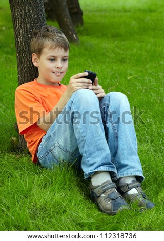 Smiling boy in orange t-shirt sits on grass under tree in park and plays with electronic game