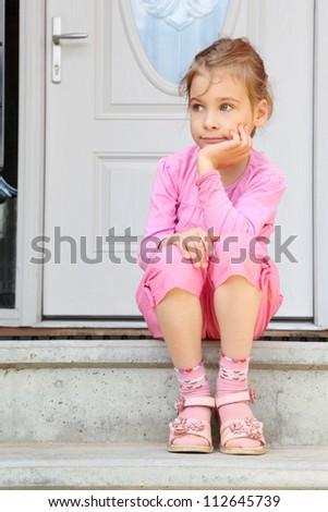Little girl sits on stairs near white door, smiles frigidly and looks away.